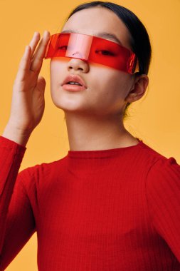 Parlak kırmızı kazaklı ve modern kırmızı yarı şeffaf vizörlü genç bir kadın stüdyo fotoğrafında canlı sarı arka planda eli gözlüklü şık bir şekilde poz veriyor..