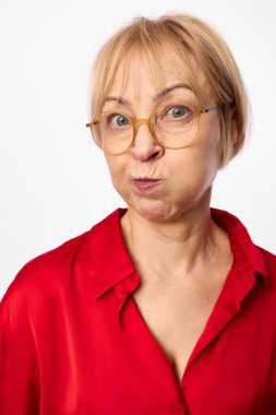 Gözlüklü, kırmızı bluzlu, neşeli yüzlü sarışın kadın. Beyaz arka planda izole edilmiş, stüdyo çekimi gelişigüzel havayı ve olgun bir kadının tuhaf yüz hareketlerini yakalıyor..