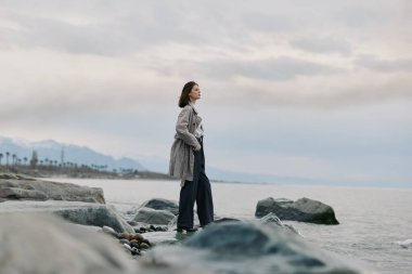 Uzun bir palto giyen düşünceli bir kadın bulutlu gökyüzü ve uzak dağlarla sakin deniz suyunun yanında kayaların üzerinde duruyor. Doğal manzara, dışarıdaki sakinliği ve yansıtıcı havayı çağrıştırıyor..