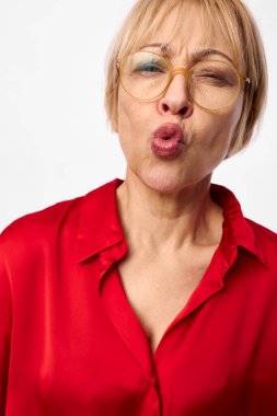 Gözlüklü, kırmızı bluzlu, büzüşmüş dudaklarıyla öpücük üfleyerek neşeli ruh halini ve mutluluğunu ifade eden olgun bir kadının yakın çekimi. Beyaz arka planda, stüdyo ışıklandırmasıyla izole edilmiş..
