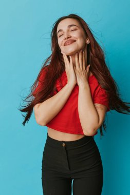 Kırmızı gömlekli, siyah pantolonlu, uzun saçlı, mavi arka planda gözleri kapalı gülümseyen kendine güvenen genç bir kadın. Rahat, mutlu, sıradan moda ve doğal ifade ile kadın portresi.