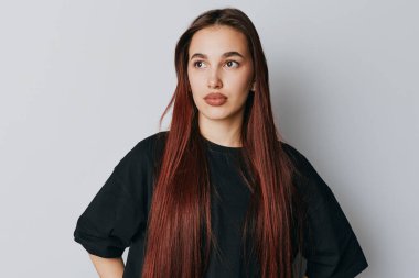 Siyah tişörtlü, uzun saçlı, elleri kalçasında arka planda poz veren genç bir kadın portresi. Kadın model, günlük kıyafet, düşünceli görünüm ve yaşam tarzı konsepti için doğal ışıklandırma..