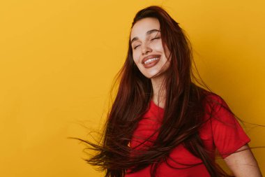 Kırmızı tişörtlü genç bir kadın. Gözleri parlak sarı duvara kapalı gülümsüyor. Dalgalı saçlı, neşeli bir kızın yakın plan portresi. Stüdyoda rahat bir ruh hali ve canlı bir rengi var..