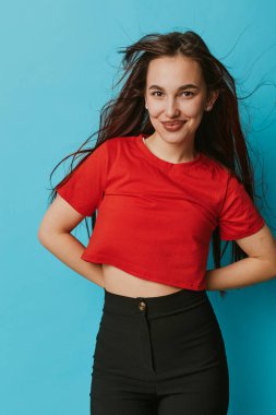 Uzun saçlı, kırmızı kısa tişörtlü, siyah pantolonlu, belinde elleri, belinde duran, mavi, pürüzsüz arka planda gülümseyen kendine güvenen bir kadın manken..