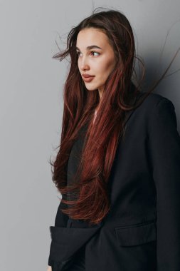 Siyah ceketli, kendine güvenen genç bir kadının portresi. Gri arka planda poz veriyor. Yumuşak ışık ve doğal ifade tarzını, zarafetini ve modern modasını yansıtıyor..