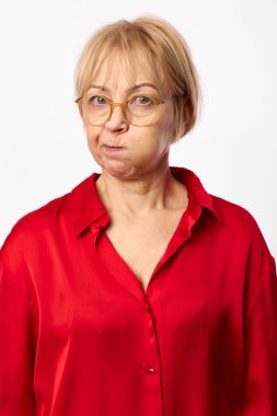 Gözlüklü, kırmızı bluzlu, yanaklı komik olgun bir kadın beyaz arka planda izole bir şekilde kameraya bakıyor. Şakacı ifade anlayışı, mizah ve samimi portre havası..