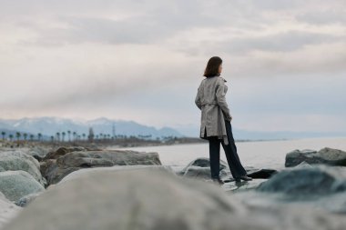 Yağmurluk giyen kadın bulutlu bir günde kayalık sahilden denize bakıyor. Arka planda dağlar ve palmiye ağaçları olan kıyı manzarası yalnızlığı, doğayı ve açık hava yaşam tarzını gösteriyor..