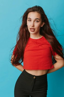 Kendine güvenen, uzun siyah saçlı, kırmızı bluzlu ve mavi arka planda duran siyah pantolonlu genç bir manken. Günlük kıyafet, stüdyo çekimi, doğal poz ve doğrudan bakış..