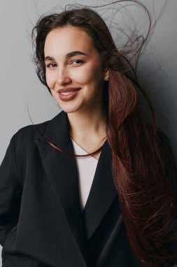 Siyah ceketli, beyaz gömlekli, gülümseyerek poz veren dalgalı saçlı genç bir kadın. Tarafsız arka planda doğal görünüşü ve küpeleri olan çekici bir kadının stüdyo portresi.