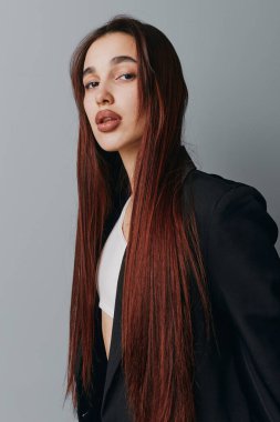 Uzun kahverengi saçlı, doğal makyajlı, siyah ceketli ve beyaz gömlekli, tarafsız gri arka planda duran kendine güvenen genç bir kadın modelin portresi..