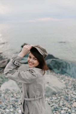 Bej ceket ve şapka giyen mutlu genç bir kadın çakıl taşı sahilinde duruyor. Sakin deniz ve bulutlu bir arka plan. Sonbahar stili, doğa ve açık hava portresi kavramı..