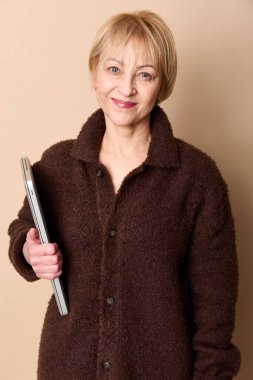 Kahverengi ceketli, kendine güvenen olgun bir kadın dizüstü bilgisayarı tutuyor ve bej arka planda gülümsüyor. Teknoloji kavramı, gelişigüzel moda ve orta yaşlı kadın portresi..