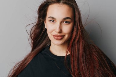 Siyah tişörtlü, düz saçlı, gülümseyen genç bir kadın tarafsız gri arka plana karşı poz veriyor. Doğal görünüşlü yakın plan portre ve moda ve sağlık için pozitif ruh hali..