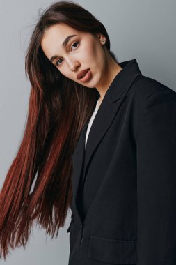 Siyah ceketli, düz saçlı bir kadın gri duvarın önünde dikiliyor. Çekici bir kadın portresi. Makyajı yumuşak ve sakin bir yüzü var..