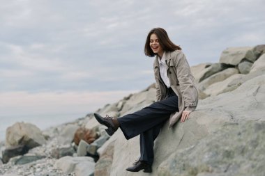 Sıradan ceket ve pantolon giyen mutlu genç bir kadın okyanusun yakınındaki kayalarda oturuyor, neşeyle gülümsüyor. Bulutlu gökyüzü ve doğal kayalık sahil huzurlu bir açık hava ortamı sağlar..