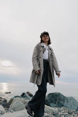 Şık palto ve şapka giyen kendine güvenen genç bir kadın bulutlu gökyüzünün altında sakin deniz kıyısında kayalık kıyılarda geziniyor. Doğal elementler ve barışçıl atmosferli açık hava yaşam tarzı ve günlük moda kavramı.