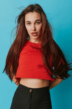 Mavi arkaplanın önünde duran uzun saçlı, kırmızı bluzlu ve siyah pantolonlu genç bir kadın portresi. Şık kıyafetler, rahat duruş ve modern moda imajına doğrudan bakış..