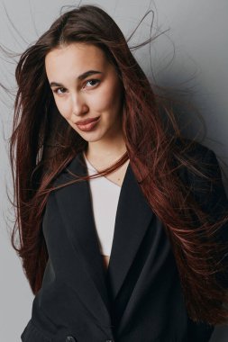 Siyah ceketli, beyaz gömlekli, dalgalı saçlı, kendine güvenen genç bir kadın. Doğal görünüşü ve ince gülüşü olan bir kadın model portrede düz gri bir duvara karşı duruyor..