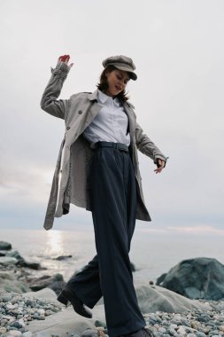 Tüylü palto, kep ve bol pantolon giyen şık bir kadın okyanus kıyısında çakıl taşı pozu veriyor. Yumuşak doğal ışık, bulutlu havada gündelik şık tarzı ve sakin kıyı atmosferini vurgular.