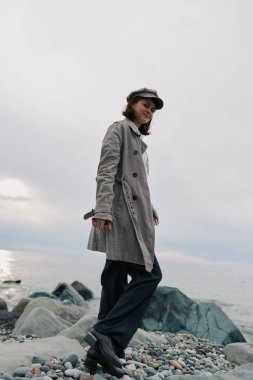 Yağmurluk ve şapka giymiş bir kadın bulutlu bir günde okyanus kıyısında çakıl taşları üzerinde duruyor. Günlük kıyafet kavramı, sahil yürüyüşü, doğa ve hafif ışıklandırmalı serin hava..