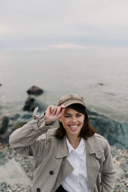 Açık renk palto ve şapka giyen mutlu genç bir kadın kayalık sahilde huzurlu su kenarında poz veriyor. O, şapkasını düzeltirken parlak bir şekilde gülümser. Seyahat, doğa ve günlük moda kavramı.