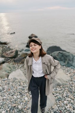 Ceket ve şapka giyen gülümseyen kız arka planda deniz ve kayalarla çakıl taşı sahilinde poz veriyor. Günlük kıyafet kavramı, doğa yürüyüşü, rahatlatıcı ruh hali ve barışçıl açık hava ortamı..
