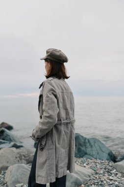 Gri montlu ve desenli şapkalı bir kadının kayalık sahilden huzurlu okyanusa bakışının yan görüntüsü. Yansıma havası, doğa ve mevsimsel stil..