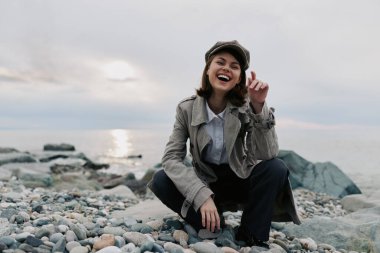 Yağmurluk ve şapka giyen mutlu genç bir kadın bulutlu gökyüzünün altında sakin denizin yanındaki kayalık sahilde çömelmiş duruyor. Moda anlayışı, neşe, açık hava yaşam tarzı, doğal güzellik ve deniz kenarında dinlenme havası..