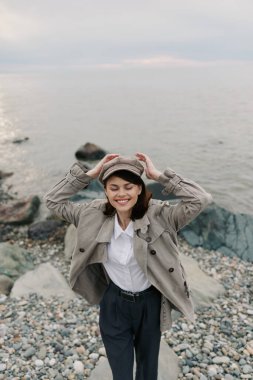 Ekose ceket giyen ve şapkalı genç bir kadın kayalık bir sahilde duruyor. Gözleri kapalı ve gülümsüyor, şapkasını meltemde tutuyor. Sonbahar stili, doğa, neşe ve açık hava yaşam tarzı kavramı.