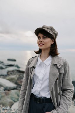 Kayalık deniz kıyısında şapkalı ve dokulu montlu genç bir kadının portresi. Sakin su ve arka planda bulutlu gökyüzü. Açık hava yaşam tarzı, mevsimsel tarz ve sükunet kavramı.