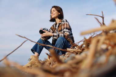 Dal ve kum arasında oturmuş, doğal ortamdaki sakin manzarayı ve huzur dolu yalnızlığı yakalayan, kameralı bir kadının sıradan dış portresi..