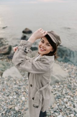 Trençkotlu ve kareli şapkalı mutlu kadın su kenarında taş plajda duruyor. Doğal ışık, kıyı sonbahar ortamında gülümsemesini ve rahatlamasını sağlar. Günlük yaşam tarzı kavramı.