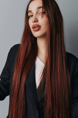 Uzun saçlı, koyu renk ceketli genç bir kadının portresi. Yüzünde hafif ışıklandırma ve pürüzsüz saç dokusuyla stüdyo çekimi, minimum makyaj ve sakin görünüm..