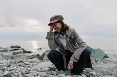 Bulutlu gökyüzünün altında, sakin deniz kenarında çakıl taşı sahilinde desenli palto ve şapka giyen gülümseyen genç bir kadın. Gündelik sonbahar kıyafetleri ve rahat pozlar neşe dolu, samimi anları dışarı taşır..