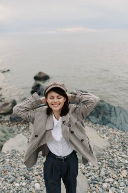 Kareli bir ceket ve uyumlu bir şapka giyen mutlu kadın kayalık bir plajda huzurlu bir anın tadını çıkarıyor. Gözleri kapalı, elleri başının arkasında, sakin bir okyanus ve bulutlu bir gökyüzü..