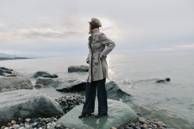 Uzun paltolu ve şapkalı kadın model okyanus kıyısındaki kayalarda duruyor. Sakin deniz ve bulutlu gökyüzü sakin bir atmosfer oluşturuyor, açık havada moda ve doğa bağlantısını vurguluyor..