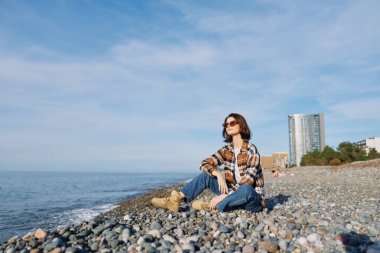 Kumsal, deniz, sahil boyunca çakıl taşları üzerinde oturan kadın güneşli gökyüzü altında rahatlamış ve manzaranın tadını çıkarıyor. Moda kıyafetleri ve sakin deniz manzarası..