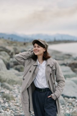 Kumaşlı palto ve şapka giyen mutlu kadın çakıl taşı sahilinde su kenarında poz veriyor. Bulutlu gökyüzü ve dağ arkaplanı, doğa fotoğrafçılığında gevşemiş ruh halini ve sonbahar tarzını vurguluyor.