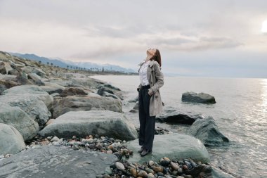 Taş plajda gözleri kapalı ve başı arkada, bulutlu gökyüzünün altında huzurlu deniz manzarasını kucaklayan rahat bir kadın. Doğa kavramı, özgürlük, farkındalık ve açık hava sakinliği..
