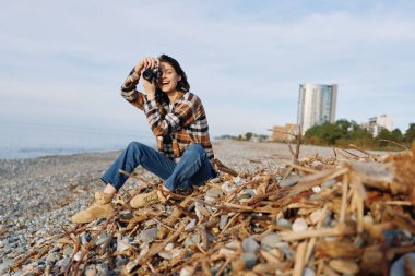 Bir plaj sahnesinde sıradan bir fotoğrafçı. Dalgakıran, kot ve ekose giyimli, odaklanmış ifade, doğal aydınlatma, samimi an, seyahat ve doğa fotoğrafçılığı..
