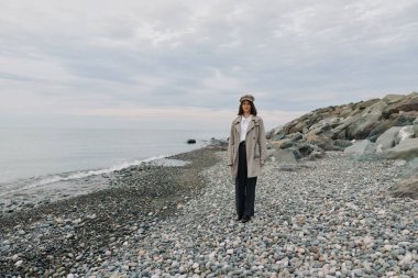 Sıcak palto ve şapkalı biri bulutlu gökyüzü ile deniz kıyısında çakıl taşında yürüyor. Yalnızlık, doğa, seyahat, sonbahar mevsimi ve doğal ortamda günlük moda kavramı..