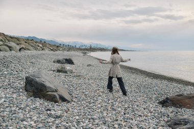 Bej bir ceket giymiş yalnız bir kadın, uzaktaki dağlarla birlikte sakin denizin kenarındaki çakıl taşlı plajda geziniyor. Ayar, sükuneti, doğayı ve iç gözlemi yansıtıyor..