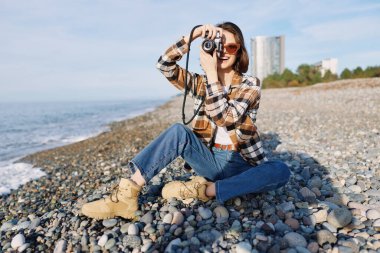Çakıl taşı sahilinde sıradan bir fotoğrafçı, objektiften çerçeve dalgalarına ve ufka odaklanıyor, kot ve ekose gömlek giyiyor, açık havada deniz fotoğrafçılığını keşfediyor..