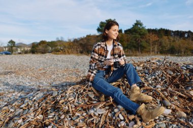 Sahilin yakınındaki yapraklardan bir yatakta dinlenen, kot, bot ve işaretli bir gömlek giyen, mavi gökyüzü ve orman manzaralı, huzurlu doğa inzivasına çekilen bir kadının olduğu sıradan bir açık hava sahnesi..