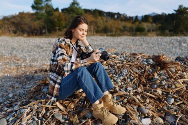 Kadın fotoğrafçı bir çakıl taşı plajında diz çöker ve çerçeveler, sonbahar ışığı kamera, ekose ceket ve kot pantolon ile samimi bir ana rehberlik eder kıyıda sakin, doğal bir açık hava sahnesi yaratır.