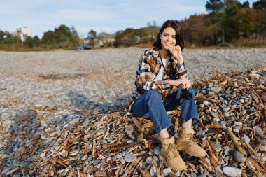 Kayalık bir plajda, ekose bluzlu, kot pantolonlu ve botlu, elinde telefon, su kenarında sakin bir anın ve uzak bir kıyı şeridindeki bir kadının sıradan portresi..