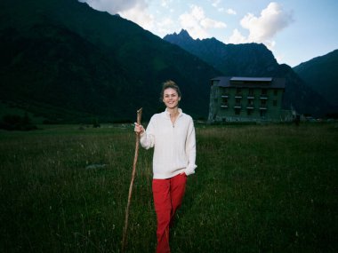 Bir vadi tarlasında asası olan gülümseyen bir kadın, engebeli dağlar ve ücra bir pansiyon, seyahat ve keşif havası, kırsal manzara ve temiz hava.