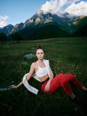 Açık hava sahnesinde çimenli bir tarlada oturan bir kadın, dağ arkası, günlük kıyafet, dingin ruh hali, doğal ışık ve geniş kırsal alan manzarası.