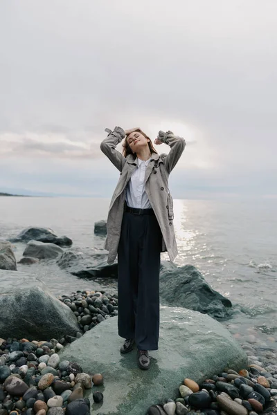 Rahat ceketli ve geniş pantolonlu bir kadın kayalık sahilde sakin deniz manzarasının tadını çıkarıyor. Bulutlu gökyüzü ve hafif ışık, rahatlatıcı havayı ve su kenarındaki doğal ortamı vurgular..