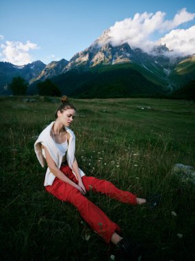 Çayırda dinlenen rahat bir kadın, dramatik bir dağ manzarası, doğa yakın, açık gökyüzü, sakin bir atmosfer, gündelik giysiler ve dağlık arazide huzur dolu bir yalnızlık.